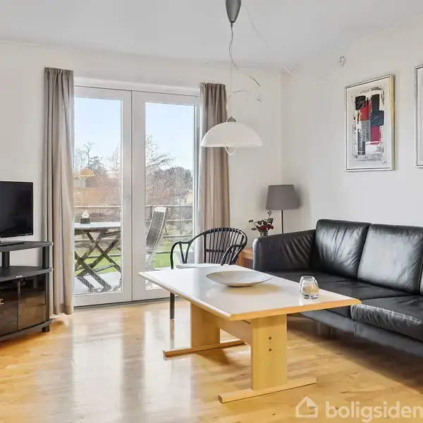 En sort sofa står ved et træbord i en stue med trægulv. Et fjernsyn er placeret på en reol, og store vinduer lukker lys ind.