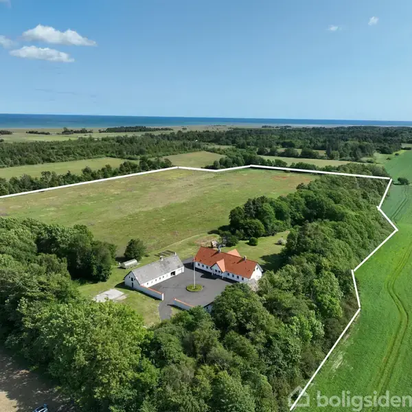 Hus med rødt tag og hvide vægge ligger stille omgivet af skov og marker. I baggrunden anes havet, og hele området er indrammet af hvide linjer.