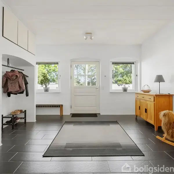 En entré med sort flisegulv. En dør flankeres af to vinduer med udsigt til grøn natur. Jakker hænger til venstre, skænk og stor spejl til højre. Genstande inkluderer en kurv og en lampe.