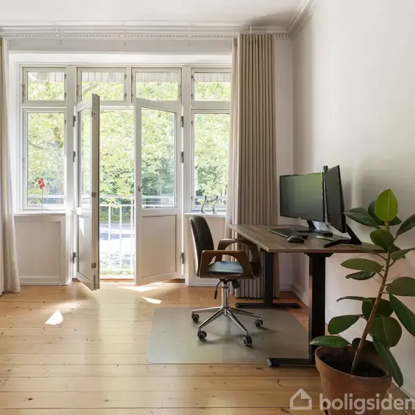 Kontorstol ved et skrivebord med computerskærm, står i et lyst rum med trægulv og åbne altandøre, omgivet af grønne planter og et vægmonteret TV.