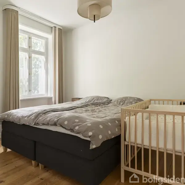En seng dækket med gråt sengetøj står ved siden af en babyseng i et lyst soveværelse med trægulv og gardiner ved vinduet.