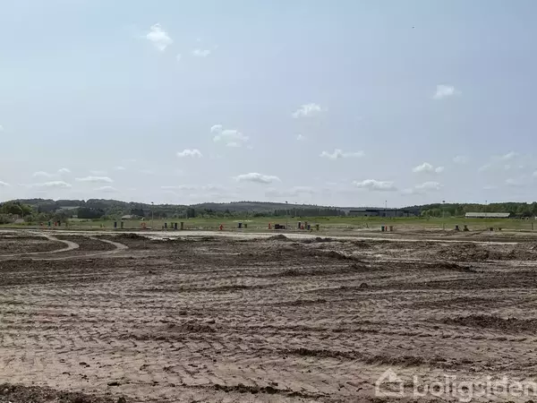 En åben, jordet mark viser spor af bulldozerdæk. I baggrunden ses spredte græsarealer og en klar, blå himmel med få skyer.