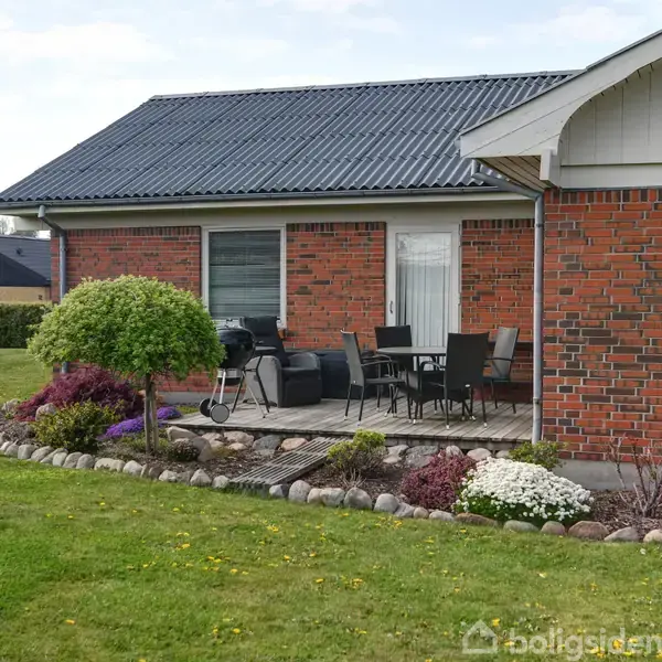En rødstenshus med terrasse, hvor der er grill og havemøbler. Det er omgivet af velplejet have med blomsterbede og klippet græs.
