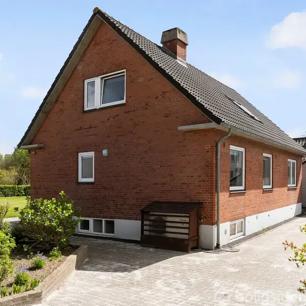 Rødt murstenshus med tag af mørke teglsten. Huset har flere vinduer og en skorsten, placeret i en belagt gårdsplads. Omkringliggende have og græsplæne fremhæver det landlige miljø.