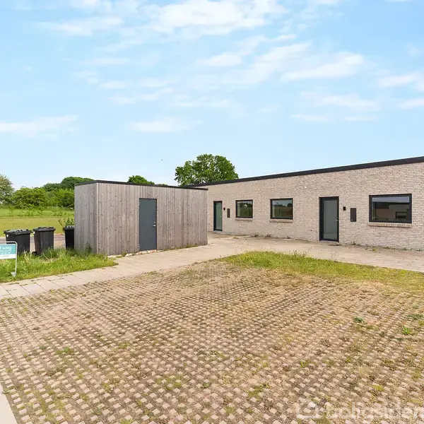 En moderne, lav murstensbygning står stille i et grønt, åbent område med parkeringsplads foran. Baggrunden indeholder græs og træer under en blå himmel.