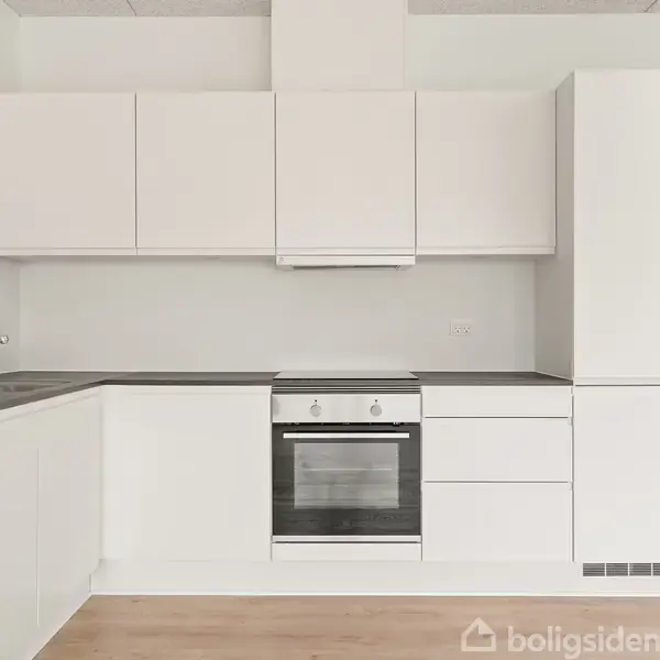 Et moderne, hvidt køkken med et indbygget komfur og ovn, placeret i en lys lejlighed. Skabene er minimalistiske, og der er en vask med en kromhane til venstre.