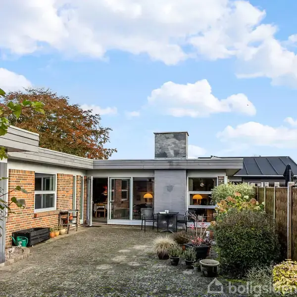 En gårdhave med blomsterkrukker og stenplads foran et murstenshus med store vinduer. Huset har en skorsten og er omgivet af buske og træer under en blå himmel med skyer.