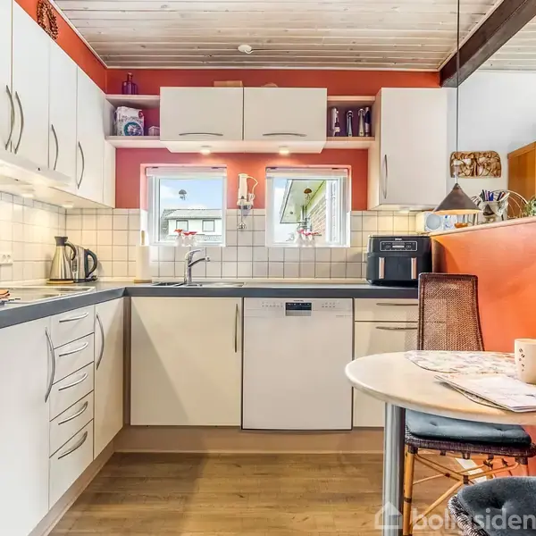 Et moderne køkken med hvide skabe og træbordplade, hvor en kaffemaskine står ved siden af en kogeplade. To små vinduer over vasken lukker lys ind i det hyggelige rum.