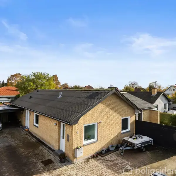 Et enkelt hus med gult murstensfacade står stille ved siden af en indkørsel, hvor en bil holder parkeret under en carport. Omgivet af en velplejet have og buske.
