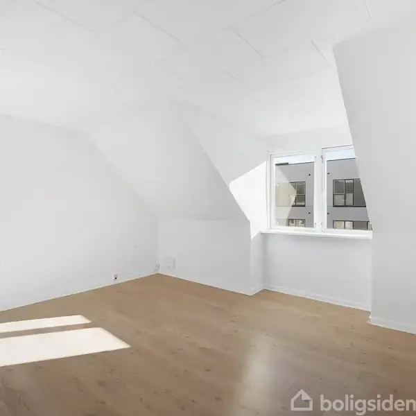 Et lyst rum med skrånende loft og trægulv, sollys strømmer ind gennem et stort, dobbeltvindue, der tilbyder udsigt til en moderne bygning udenfor.