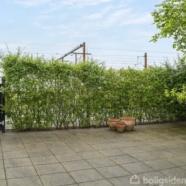 Terrasse med fliser og nogle krukker står stille. Omgivet af grønne buske, et hegn og en hvid mur. Der er blå himmel og togskinner i baggrunden.