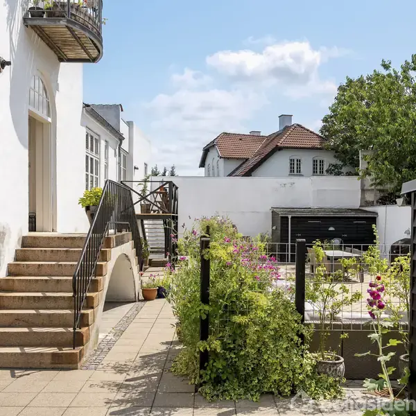 Hvid bygning med trappe, fører ud til en flisebelagt gårdhave med blomsterbede og buske. Træhegn adskiller områderne, mens andre huse og træer ses i baggrunden.