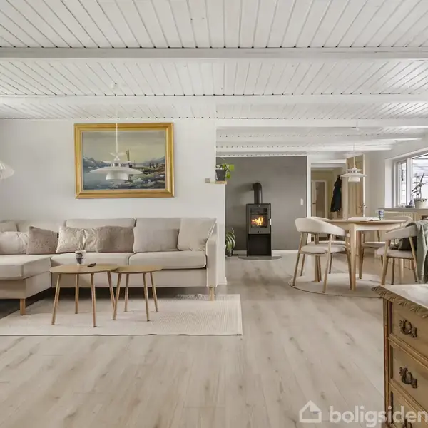Stue med beige hjørnesofa, tre små borde og maleri på væggen. I baggrunden ses spisebord med stole og en brændeovn. Lysindfald fra venstre og lyse trægulve dominerer.