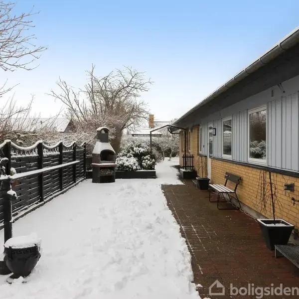 En lang, sneklædt havegang med et gult murstenshus til højre og et hegn til venstre. I baggrunden ses en udendørs pejs omgivet af sne.