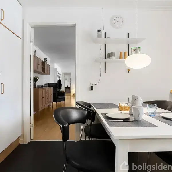 Et lyst køkken viser et spisebord med dækkede pladser, omgivet af sorte stole. En hvid lampe hænger over bordet. I baggrunden ses en åbning til en stue med møbler.