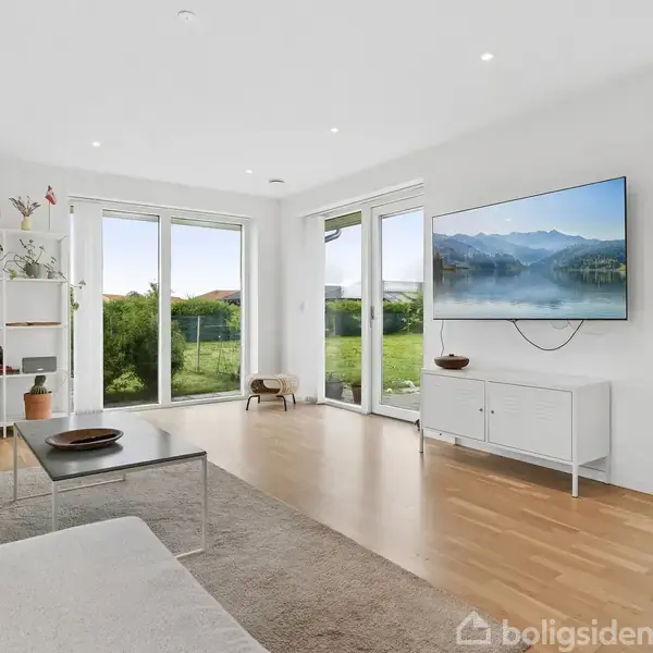 Stuen har et moderne interiør med en sofa, sofabord og reol. Et tv viser et naturbillede. Store vinduer åbner op til en have med græsplæne.