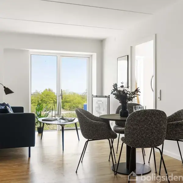 En hyggelig stue med et rundt bord og fire stole, ved siden af en sofa. Store vinduer i baggrunden lader lys ind. Der er planter og minimalistisk indretning.