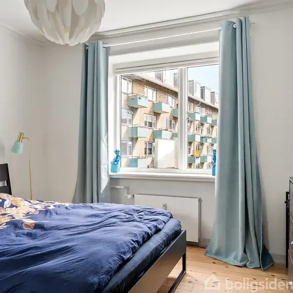 En seng med mørk træramme og blomstret sengetæppe står i et lyst soveværelse. Gardiner ved vinduet viser udsigt til en bygning med balkoner.