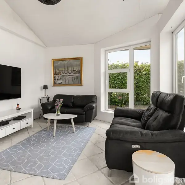 En stue med sort lædersofa og lænestol, et tv hængende på væggen over en hvid mediekonsol. Et tæppe ligger på gulvet ved siden af et rundt bord.