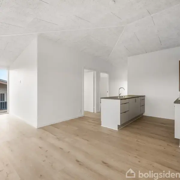 En rummelig træterrasse med glasdøre er åben mod et moderne hus med sorte træpaneler. Terrassen har udsigt over nabohuse under en klar, blå himmel.
