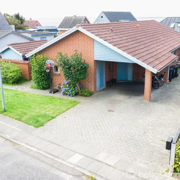 En enkel etplans murstenshus med skråt tag er beliggende i et villakvarter. Der er en flisebelagt indkørsel med en carport, og huset er omgivet af grønne buske og planter.