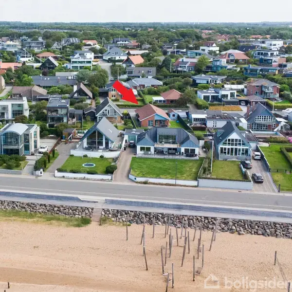 Strandbillede med moderne huse langs en vej. Græsarealer bag husene og stranden i forgrunden. En rød pil peger mod et specifikt hus i baggrunden.