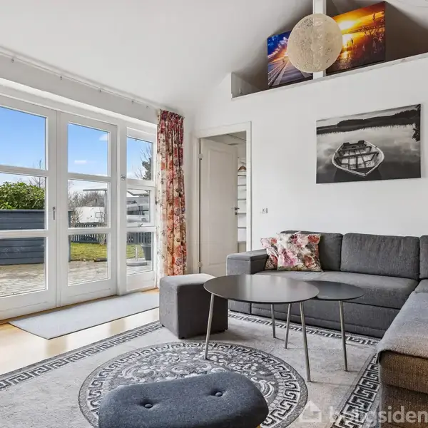 Stue med grå hjørnesofa fyldt med farvede puder, et rundt sofabord og lyse trægulve. Store glasdøre fører ud til terrasse og have. Vægkunst hænger over sofaen.