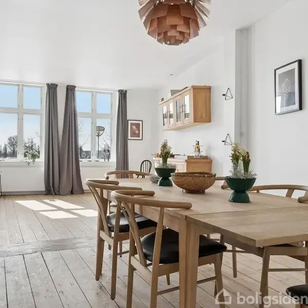 En spisestue med et træbord og stole placeret på et trægulv. Rummet er lyst med store vinduer og gardiner, dekoreret med billeder på væggen og grønne planter.