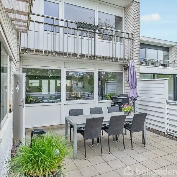 Terrasse med et spisebord og seks stole, placeret foran en moderne bygning med store vinduer. Parasol står oprejst, og plantekasser tilføjer grønne elementer i den flisebelagte gårdhave.