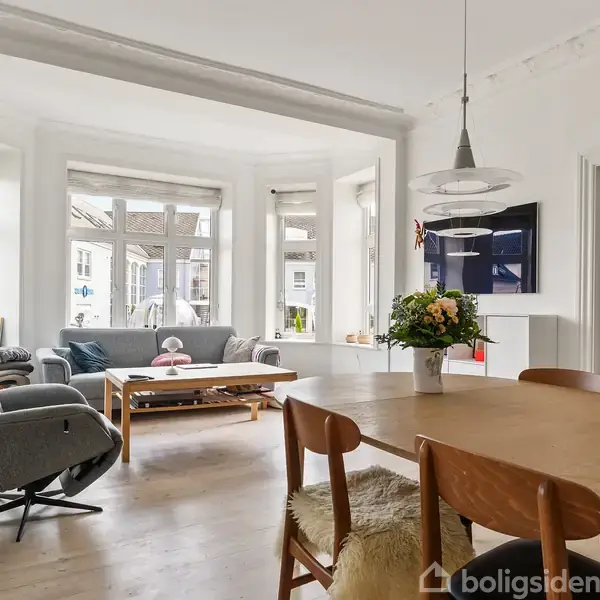 En spisestue med træbord og stole, ved siden af en opholdsstue med en sofa og lænestol. Store vinduer lader naturligt lys ind. En vase med blomster står på bordet.