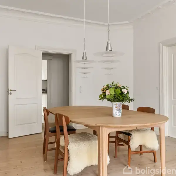 Et rundt spisebord i træ er centralt placeret med fire stole, pyntet med fåreskind, omkring. På bordet står en blomsterdekoration. Rummet har lyse vægge, og en åben dør fører til et tilstødende værelse.