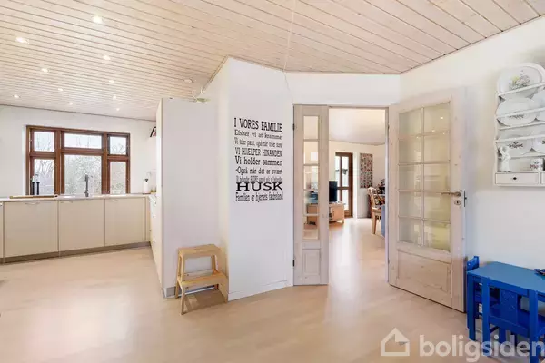 Tekst på væggen: "I VORES FAMILIE: Elsker vi med hinanden. Vi har det sjovt. Vi fejrer succeser. Vi holder sammen. Opfører os ordentligt. Husk Familien er hjemmets hjerteblod." Stue med køkken i baggrunden, lys træindretning og åbne skydedøre mellem rummene.