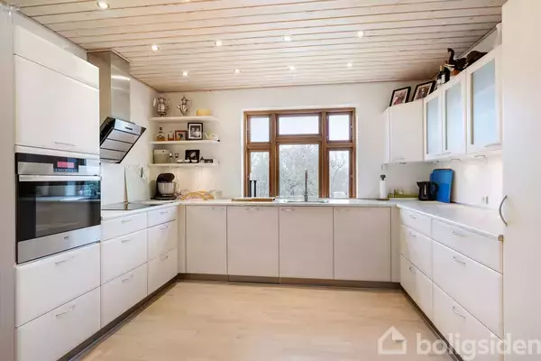 Et moderne køkken med hvide skabe og skuffer, indbygget ovn og mikroovn. Rummet er lyst, med trægulv og loft. En stor vindue viser udsigt til træer udenfor.