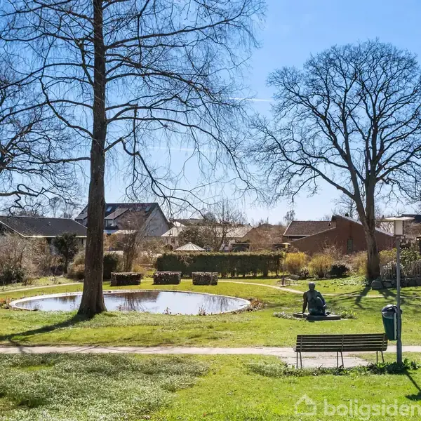 En statue sidder ved siden af en lille dam i en park med græs og træer. Bænke og en affaldsspand er placeret langs en sti, huse i baggrunden.