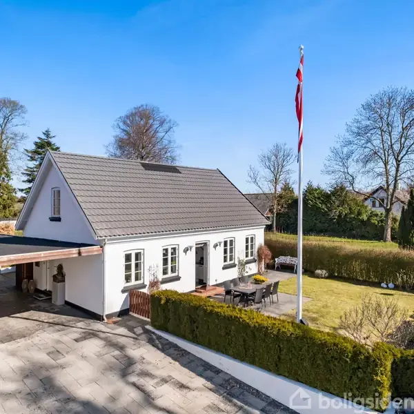 Hvidt hus med grå tagsten, omgivet af velplejet have med dansk flagstang. Til venstre, en overdækket carport. Baggrunden viser træer under en klar blå himmel.