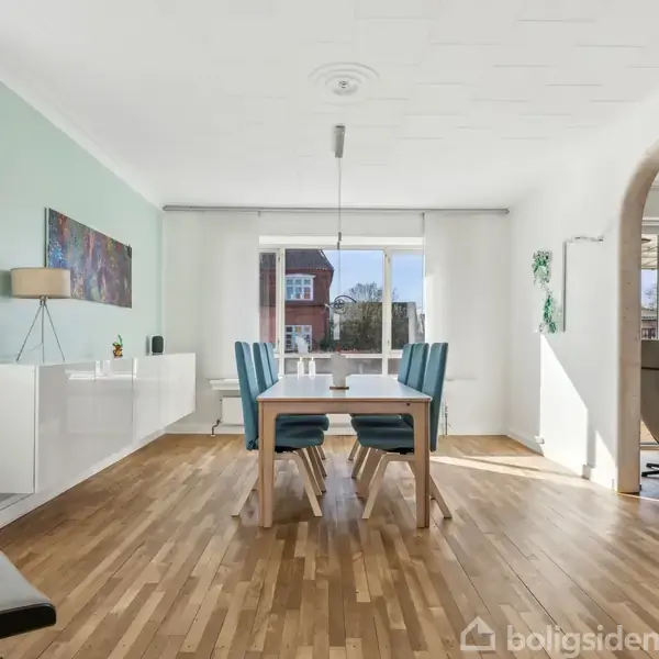 Spisebord med blå stole står på trægulv. Rundt omkring er der hvide vægge med vinduer. En bue åbner til et tilstødende rum, og dagslys strømmer ind.