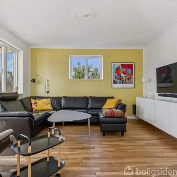 Stue med sort lædersofa, placeret L-formet mod en gul væg. Fladskærms-TV er monteret på en hvid skænk. Store vinduer til venstre lader naturligt lys ind.