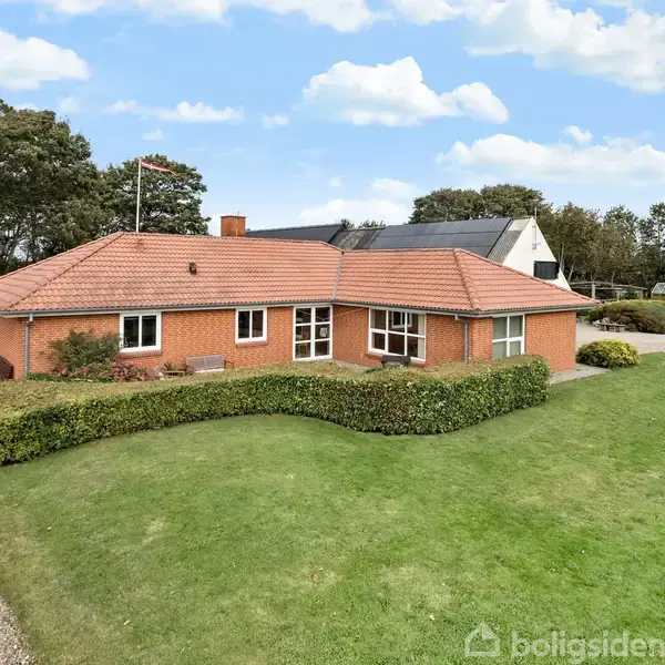 Et rødt murstenshus med rødt tegltag står på en velplejet græsplæne med en grusvej ved siden af. Hække omkranser huset i et landligt område.