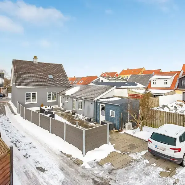 En hvid bil holder parkeret i en snedækket indkørsel foran et gråt hus med skråt tag. Omgivet af flere huse i en villakvarter under en klar blå himmel.