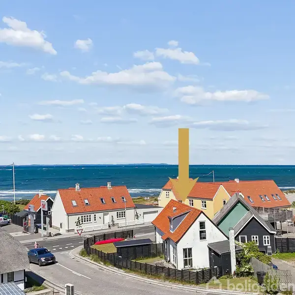 En klynge af huse med røde tage står tæt på hinanden, mens en pil peger på en specifik bygning. Havet og en blå himmel fylder baggrunden.