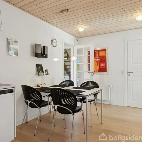 En spisestue med sort bord og fire sorte stole, omgivet af lyse trævægge og loft. Der hænger en pendellampe over bordet og en rød kunstplakat på væggen.