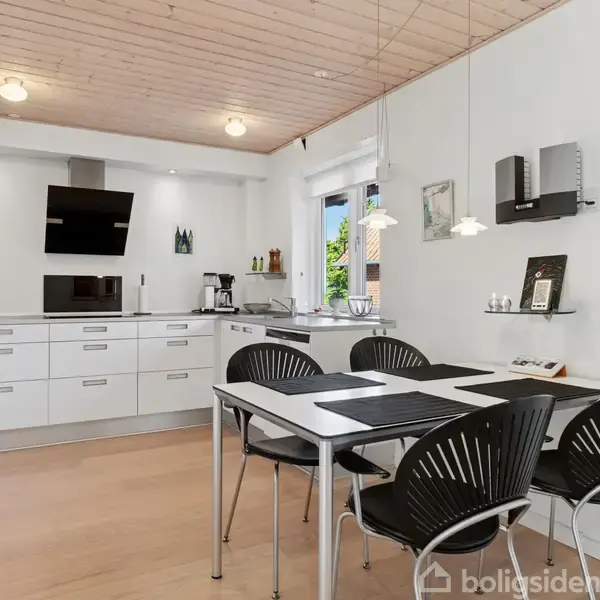 Et moderne køkken med hvide skabe og en indbygget ovn. Et sort komfur og en emhætte er centralt placeret. Et sort spisebord med fire stole står i forgrunden. En vægur hænger på væggen.