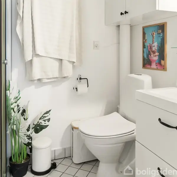 Toilet står ved siden af et håndvaskskab under et spejl. En lille plante og toiletrulleholder ses, mens et hvidt håndklæde hænger tæt ved. En plakat hænger på væggen.
