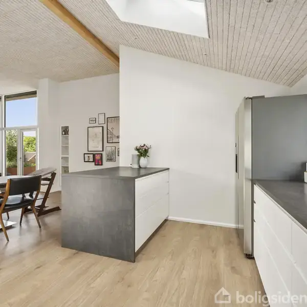 Et moderne køkken med en grå køkkenø; på bordet står blomster. I baggrunden ses en spisestue med stort vindue, træmøbler og et loft med trælister.