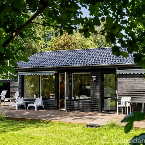 Sort træhus står stille omgivet af grønne træer og en velplejet græsplæne. En åben terrasse med hvide stole under en markise skaber en hyggelig udendørs atmosfære.