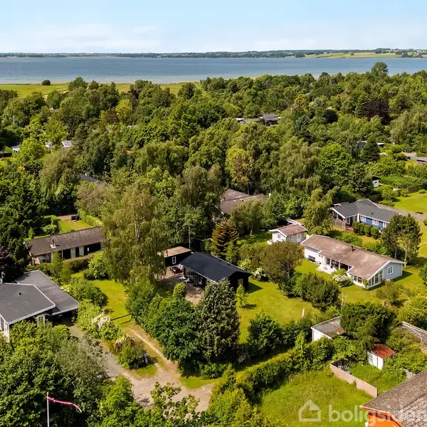 Huse er spredt ud i et frodigt, grønt område med træer og haver, beliggende ved en sø eller kystlinje i baggrunden under en blå himmel.