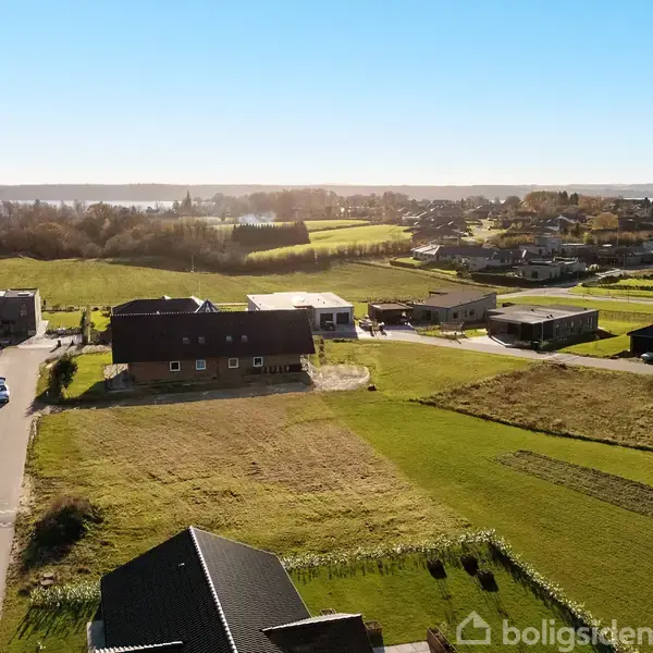 Landskab udgør grønne marker og spredte huse i en landsby nær vand. Biler holder parkeret langs en vej. Sollys oplyser området, der er omkranset af skov i horisonten.