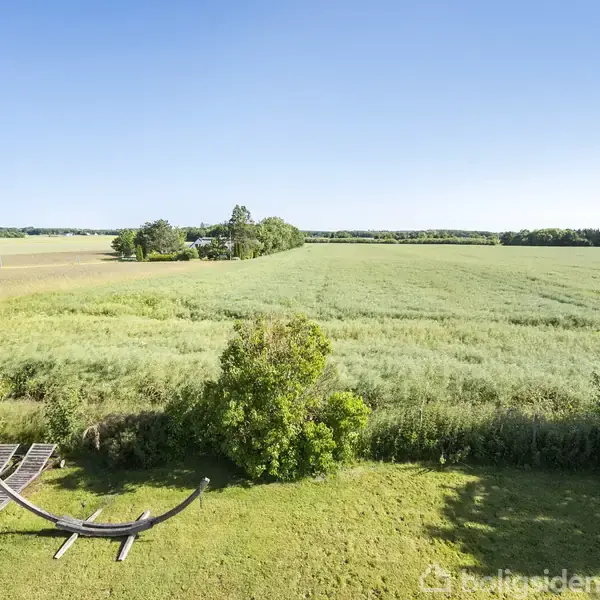 Bindingsværkshus står stille med dets stråtag blandt grønne træer og marker. To biler er parkeret foran, og flaget vajer ved siden af en pavillon i haven.