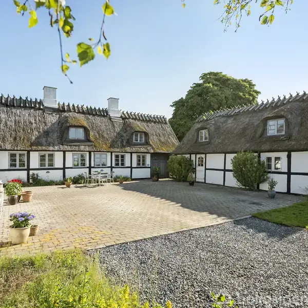 Stråtækt bindingsværkshus omkranser en brostensbelagt gårdsplads, omgivet af grønne planter. Sollys belyser gårdspladsen, og en blå himmel fuldender den idylliske atmosfære.