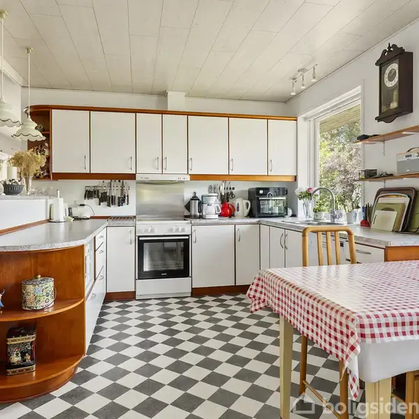 Et familie-køkken med hvidt og træfarvet interiør. Et spisebord med rødternet dug står ved siden af et skakternet gulv. Store vinduer giver udsigt og dagslys.